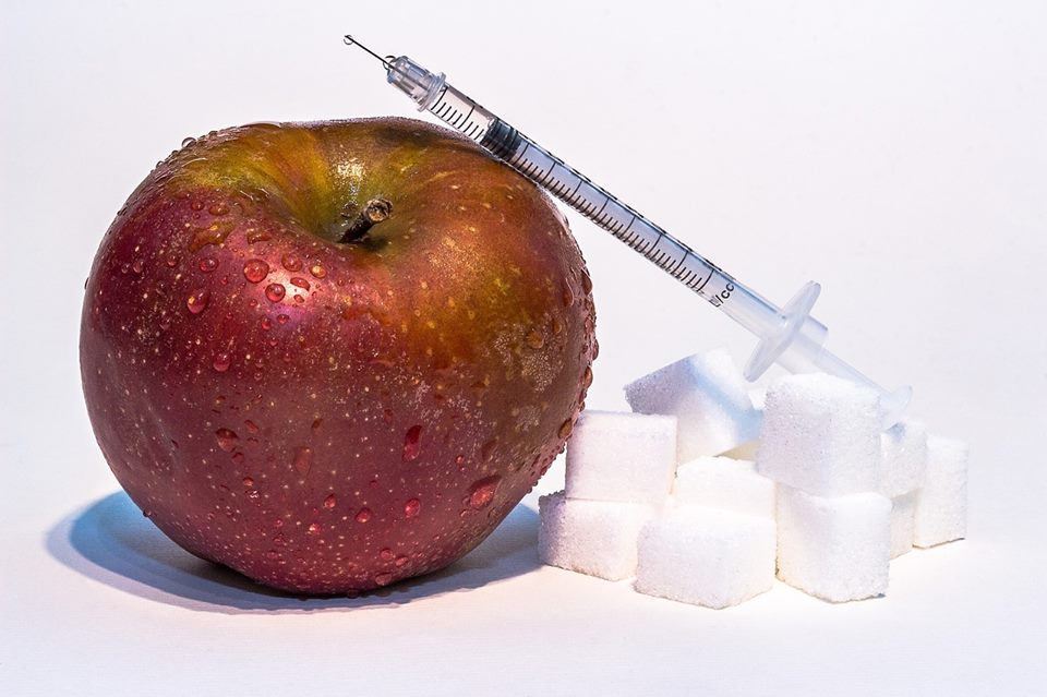 Zdjęcie jabłka, z boku strzykawka i kostki cukru. Ma wydźwięk symboliczy dotyczący cukrzycy. 