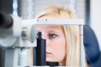 Zdjęcie badania oka na lampie szczelinowej. Widać młodą kobietę o blond włosach trzymającą głowę na lampie szczelinowej. 